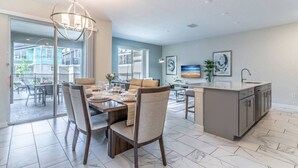 Une cuisine moderne avec un coin repas, comprenant un grand îlot central, un éclairage suspendu et une vue sur une terrasse extérieure accessible par des portes vitrées.