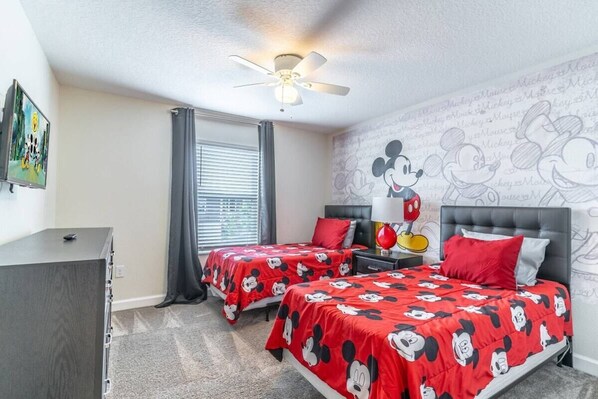 A hotel room with two beds, a ceiling fan, a nightstand with a lamp, and Mickey Mouse themed wallpaper.