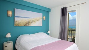 A bedroom with a bed, a painting of a beach, and a view of the sea through a window. - Centru : Appart lumineux vue mer, 5 min à pied de la plage et du centre (Calvi)