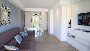 A modern living room with a sofa, a dining table, and a TV mounted on the wall. - Centru : Appart lumineux vue mer, 5 min à pied de la plage et du centre (Calvi)