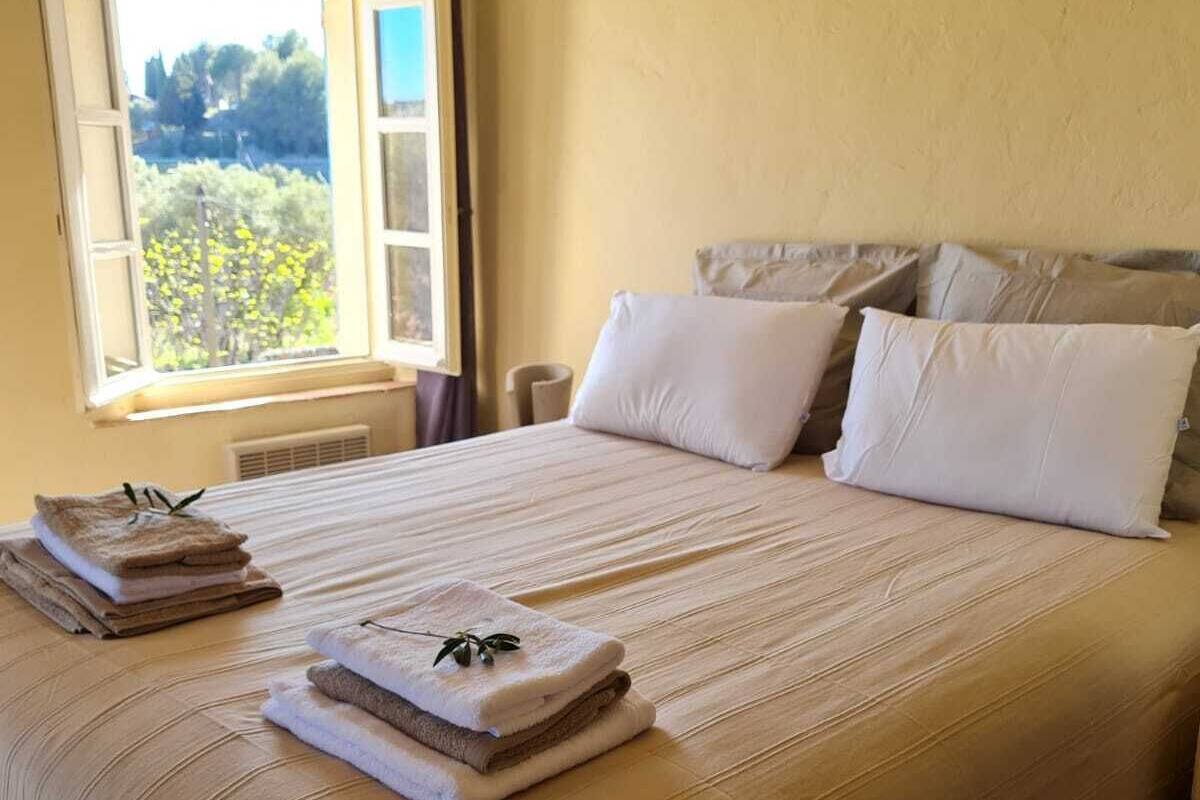 A neatly made bed with pillows and folded towels, a window with curtains, and a view of greenery outside.