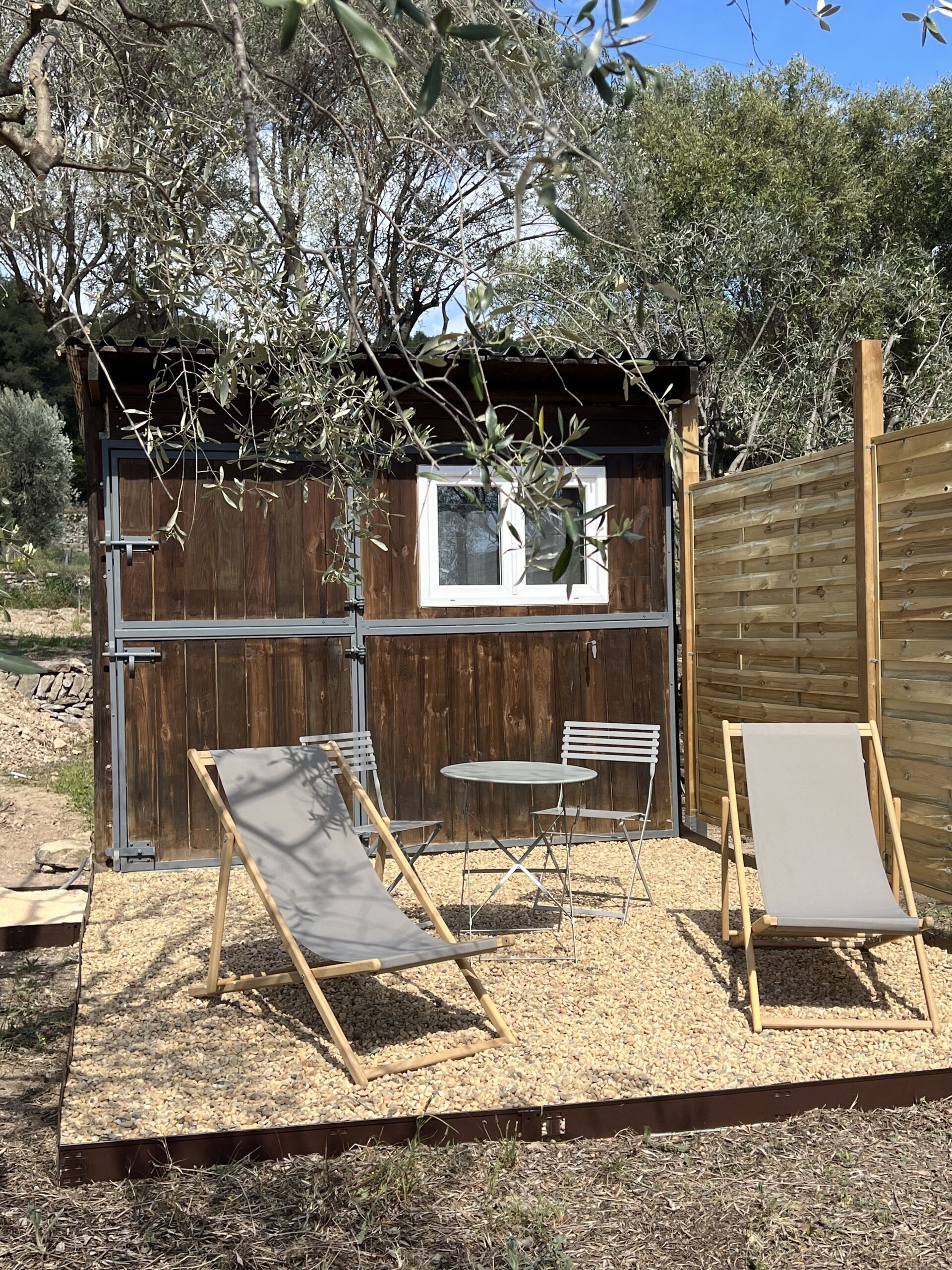 Un espacio de estar al aire libre con sillas reclinables y una mesa, rodeado por una valla de madera y olivos.