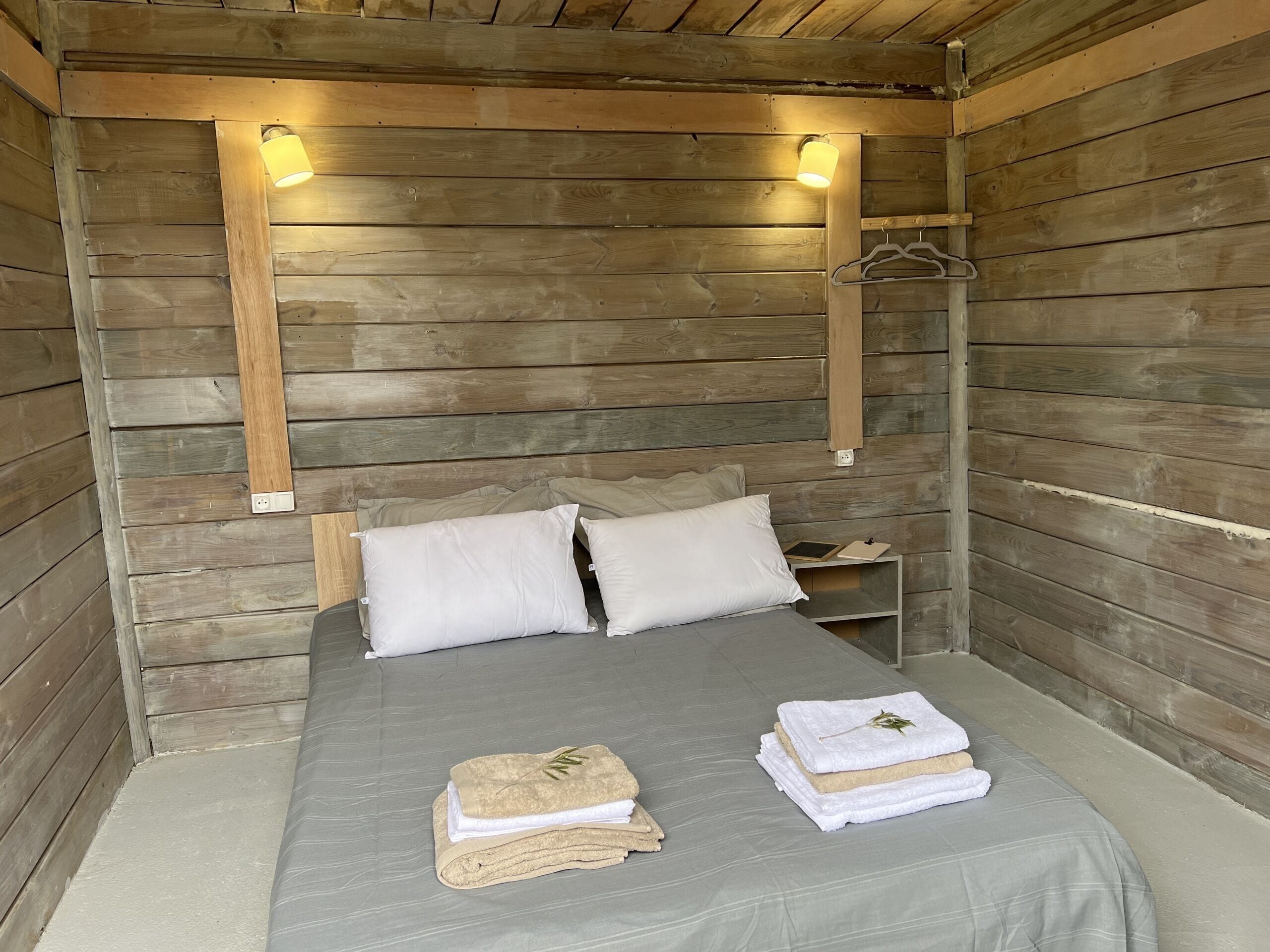 Una habitació tipus cabana de fusta, amb un llit, dues làmpades de nit, una petita taula de nit i una perxa per a abrics.