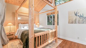 Un lit à baldaquin en bois, recouvert d’une couverture verte, une table de chevet avec une lampe, une fenêtre donnant sur un paysage verdoyant et un tapis sur le sol.