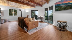 Un salon spacieux avec un plafond en bois, un canapé, une petite table d’appoint ronde et une grande fenêtre donnant sur la forêt.