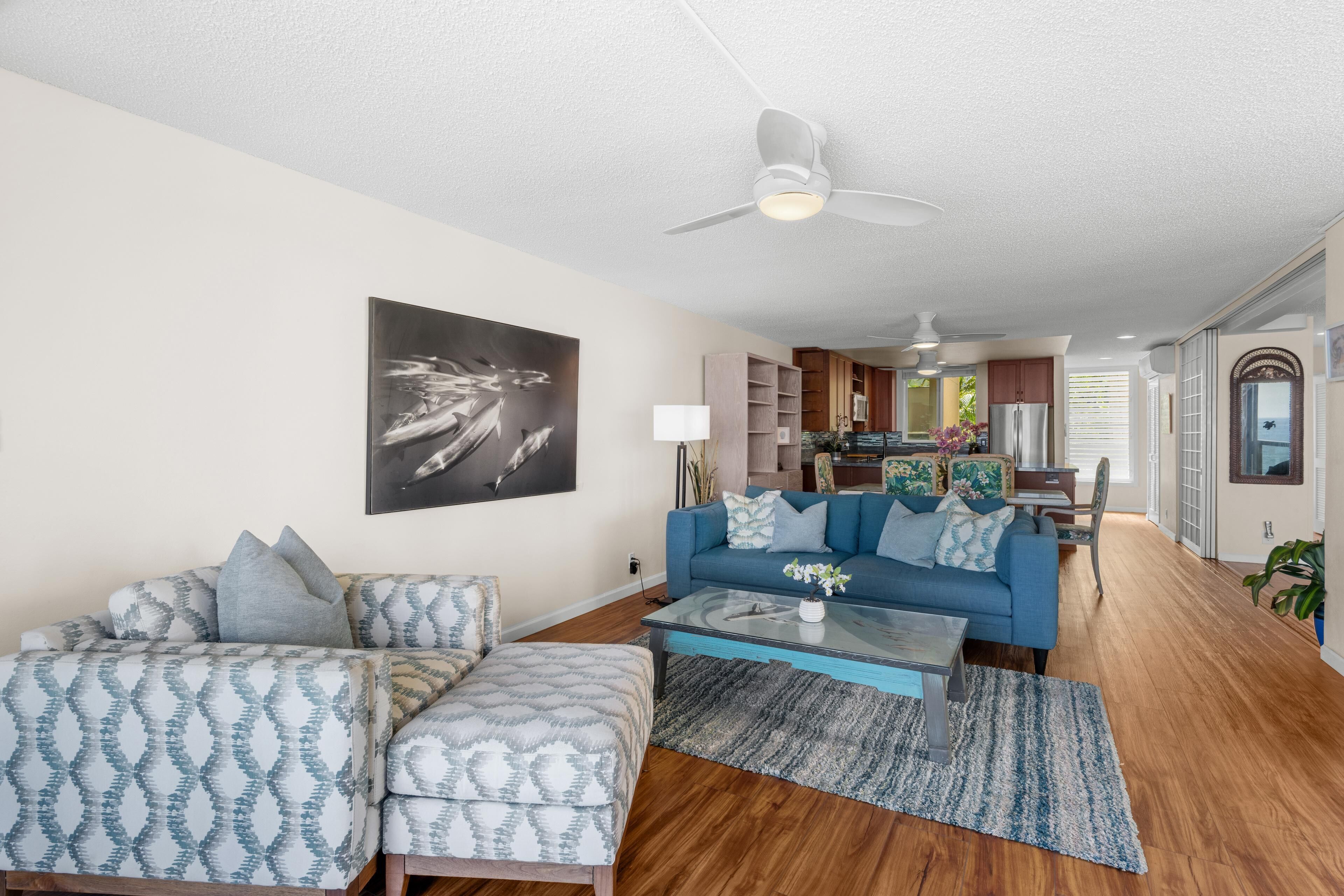 A living room with a sofa, ottoman, and coffee table, a ceiling fan, and a kitchen in the background.