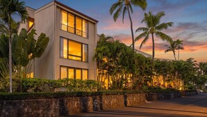 A modern two-story building with large windows, surrounded by lush tropical vegetation and a stone wall.