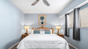 Ein Schlafzimmer mit einem Bett, zwei Nachttischen mit Lampen, einem Deckenventilator, einem gerahmten Bild an der Wand und einem Fenster mit Jalousien.