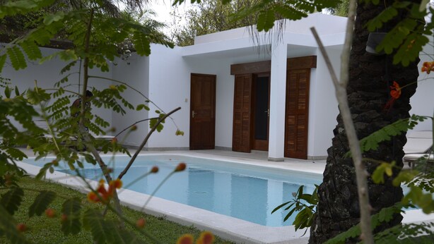A poolside area with a white building, wooden doors, and a stone pillar. - Villa Citronsverts (Cap skirring)