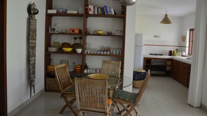 A compact kitchen with a dining area, wooden chairs, a glass table, and a bookshelf filled with books and kitchenware. - Villa Citronsverts (Cap skirring)