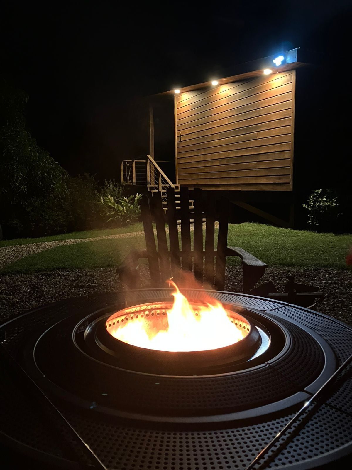 A circular fire pit with flames, surrounded by a metal grill, set on a paved area with a wooden deck structure in the background.
