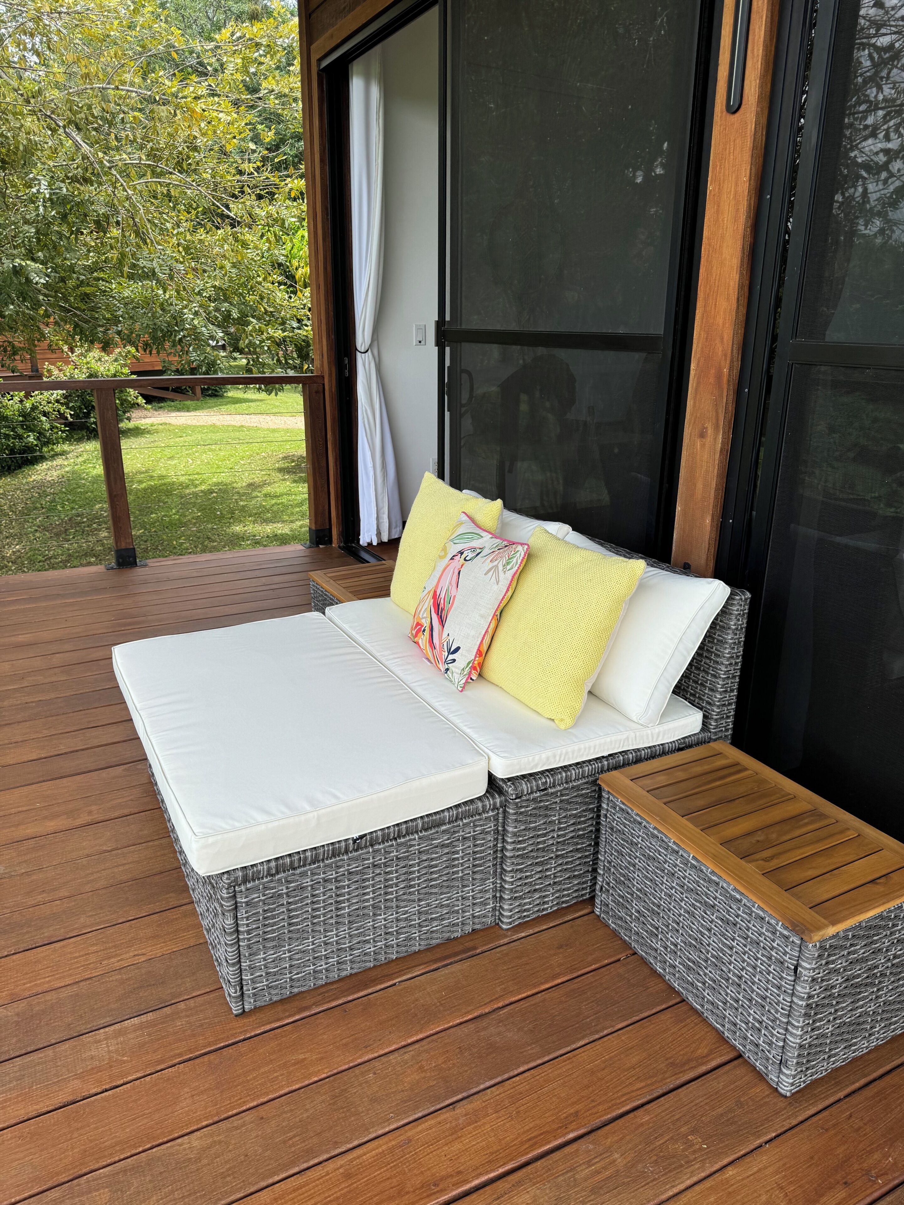 A wicker sofa with a white cushion and two pillows, one yellow and one with a floral pattern, on a wooden deck.