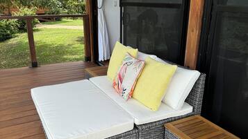 A wicker sofa with a white cushion and two pillows, one yellow and one with a floral pattern, on a wooden deck.