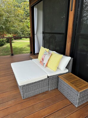 Ein Korbsessel mit weißem Kissen und zwei Zierkissen, eines gelb und eines mit Blumenmuster, auf einer Holzterrasse.