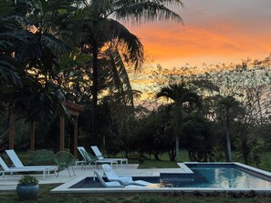 A swimming pool with lounge chairs and a wooden pergola, surrounded by lush greenery and palm trees. - Cabana 1 -Modern amenities cabin, on the banks of the Belize River (Santa Elena)