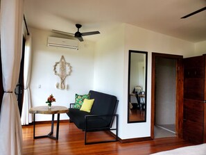 A room with a black sofa, a small round table with a plant, a ceiling fan, an air conditioning unit, and a large mirror.