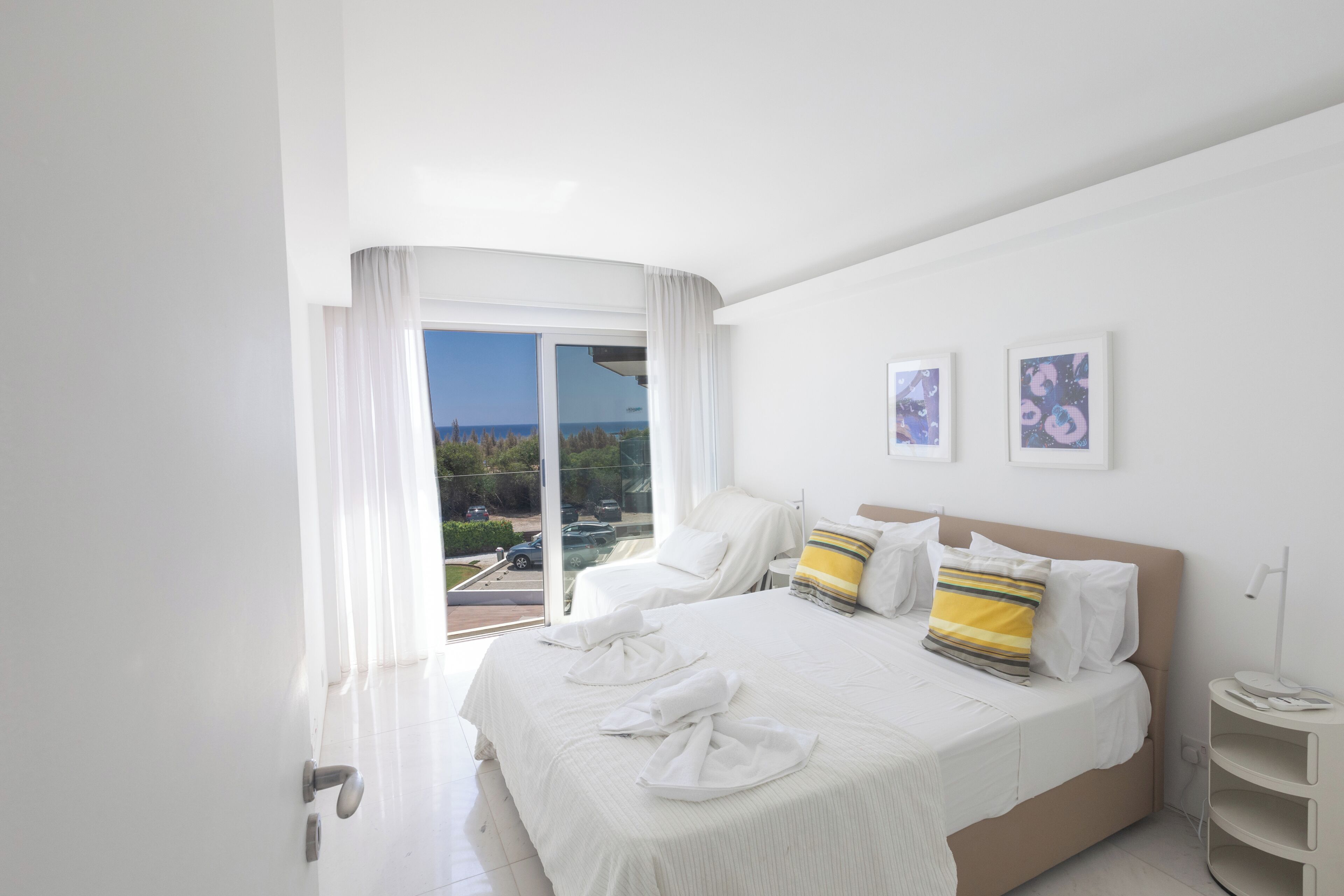 A hotel room with a large bed, a bedside table, a lamp, and a view of the outdoors.