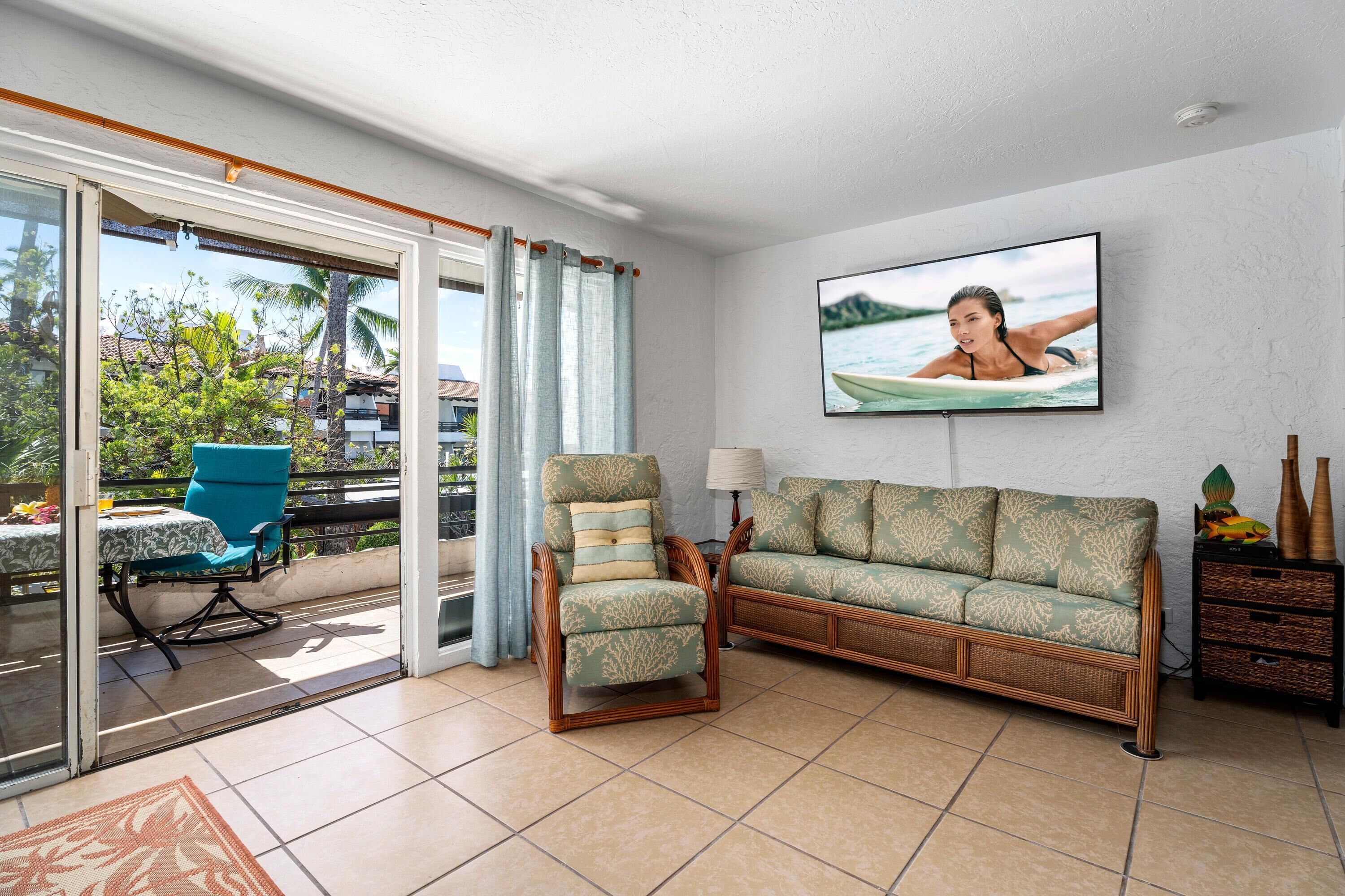 A living room with a sofa, chair, TV, and a balcony with a view of palm trees.