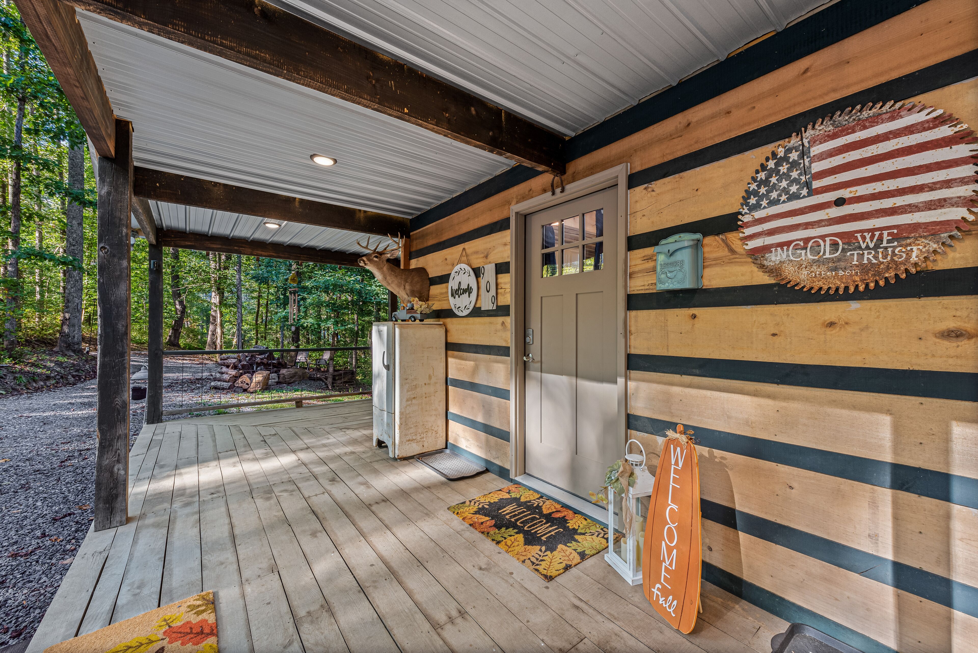Eine Holzhütte mit verdeckter Veranda, einer Fußmatte und einem Schild mit der Aufschrift 'WELCOME'. Eine rustikale amerikanische Flaggen-Dekoration ist ebenfalls zu sehen.
