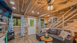 A cozy cabin interior with a wooden ceiling, a brown leather sofa, a round wooden table, and a staircase.