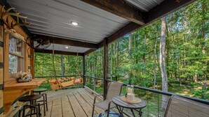 A wooden deck with a table and chairs, a hanging fan, and a large window overlooking a forest.