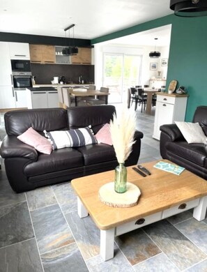 A modern living room with a black leather sofa, a wooden coffee table with a vase, and a kitchen in the background. - Nouveauté Plouescat, Grande Maison vue mer Pour 8 Personnes (Plouescat)