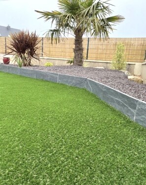 A landscaped garden with a palm tree, a small shrub, and a stone retaining wall. - Nouveauté Plouescat, Grande Maison vue mer Pour 8 Personnes (Plouescat)