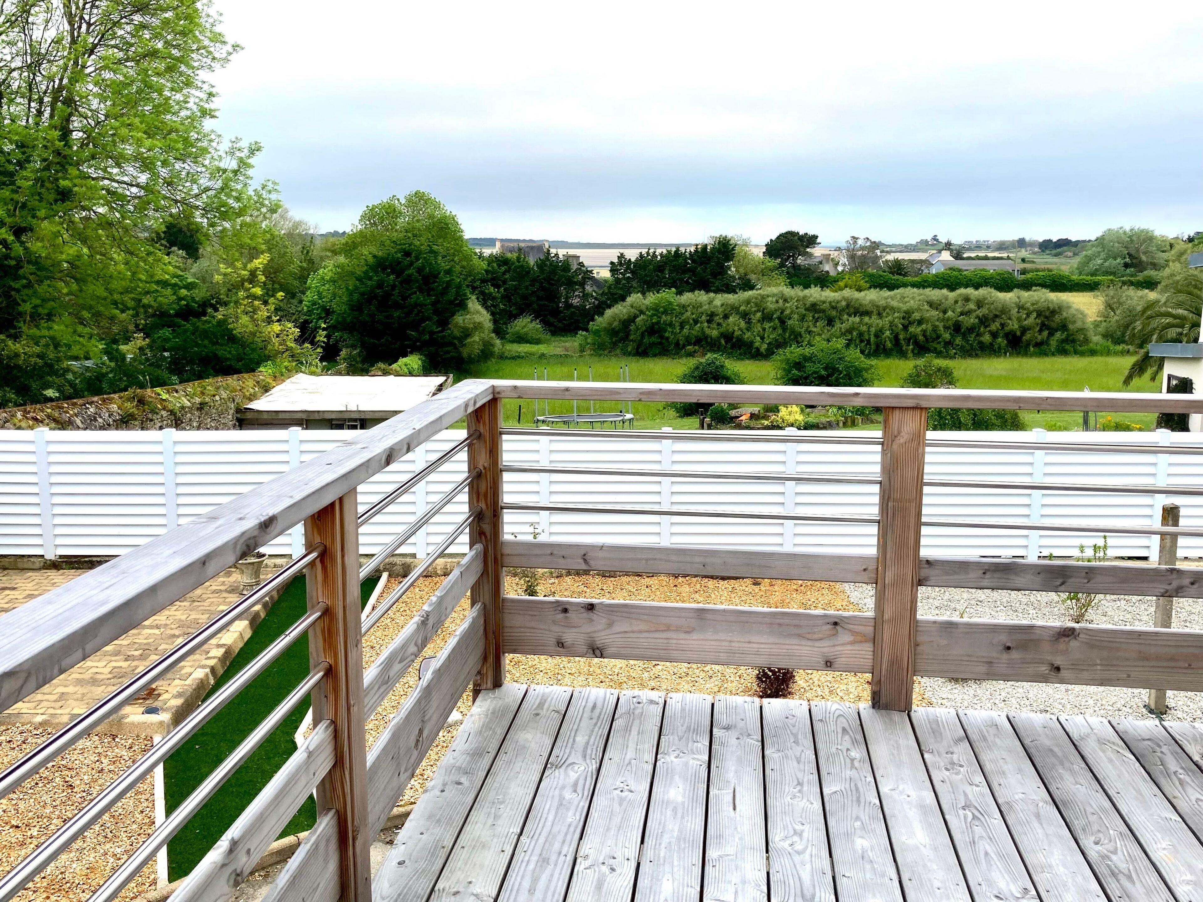 Una terraza de madera con barandilla de metal que da a un paisaje verde con árboles y una vista distante de edificios.