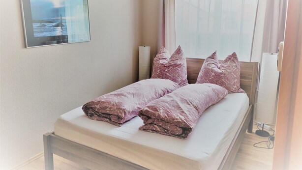 A neatly made bed with a patterned blanket and pillows, a framed picture on the wall, and a window with curtains.