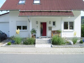 A two-story house with a red-tiled roof, a front door, and a small porch with steps leading to the entrance.