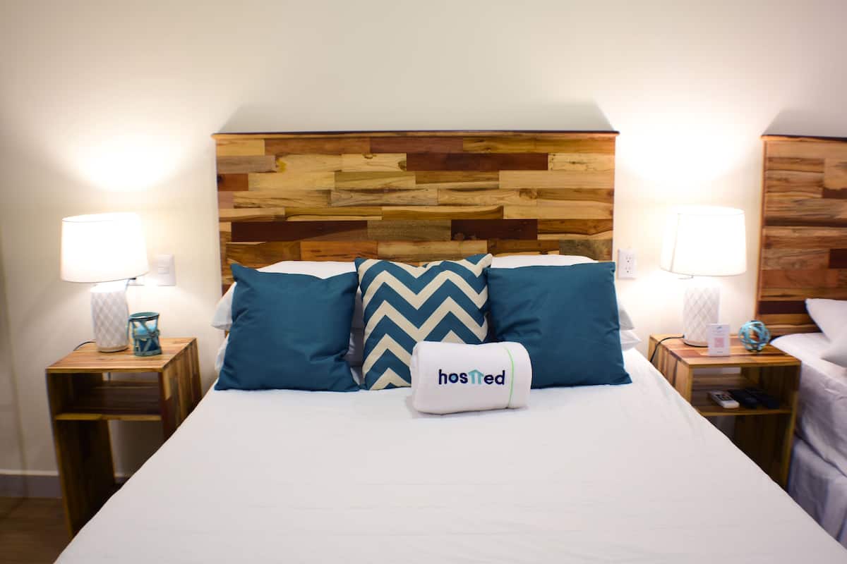 A neatly made bed with a wooden headboard, two bedside tables with lamps, and a pillow with the word "hosted" on it.