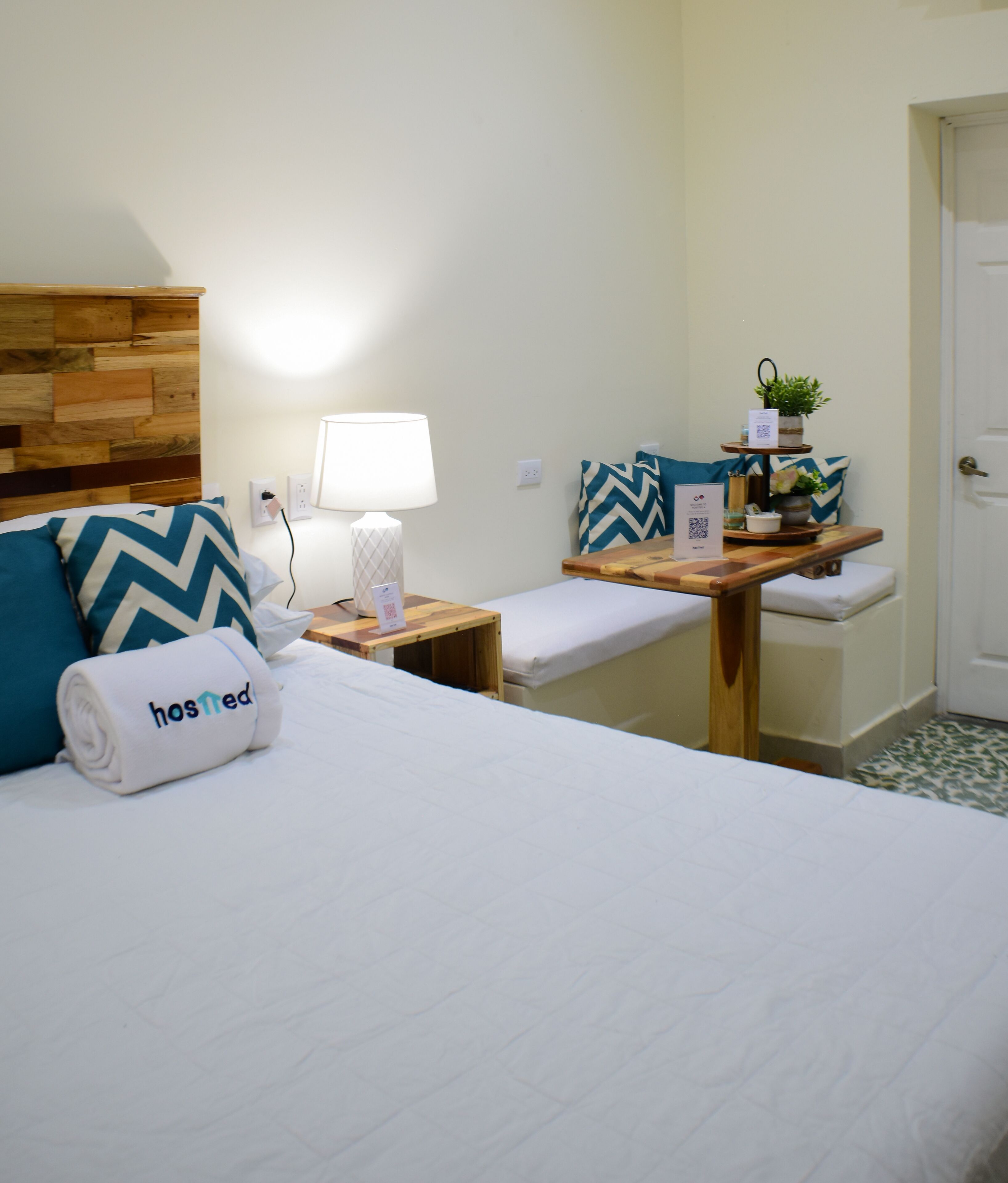 A hotel room with a neatly made bed, a bedside table with a lamp, a small shelf with decorative items, and a bench with pillows.