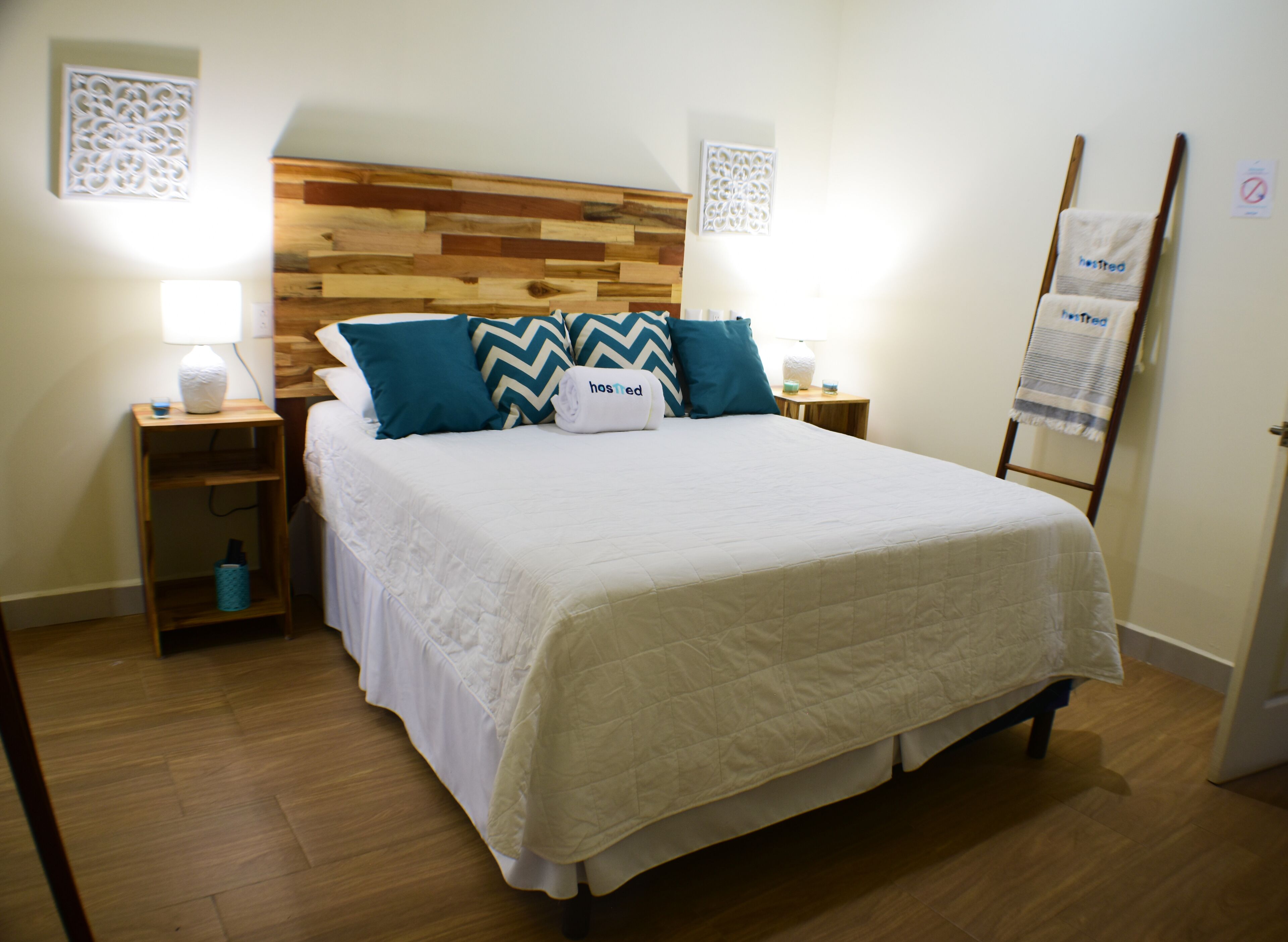 Ein ordentlich bezogenes Bett mit weißer Daunendecke und blaue sowie beige Kissen, ein Holz-Kopfschild, Nachttische mit Lampen und ein Holzboden.