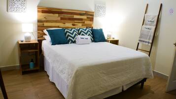 Ein ordentlich bezogenes Bett mit weißer Daunendecke und blaue sowie beige Kissen, ein Holz-Kopfschild, Nachttische mit Lampen und ein Holzboden.