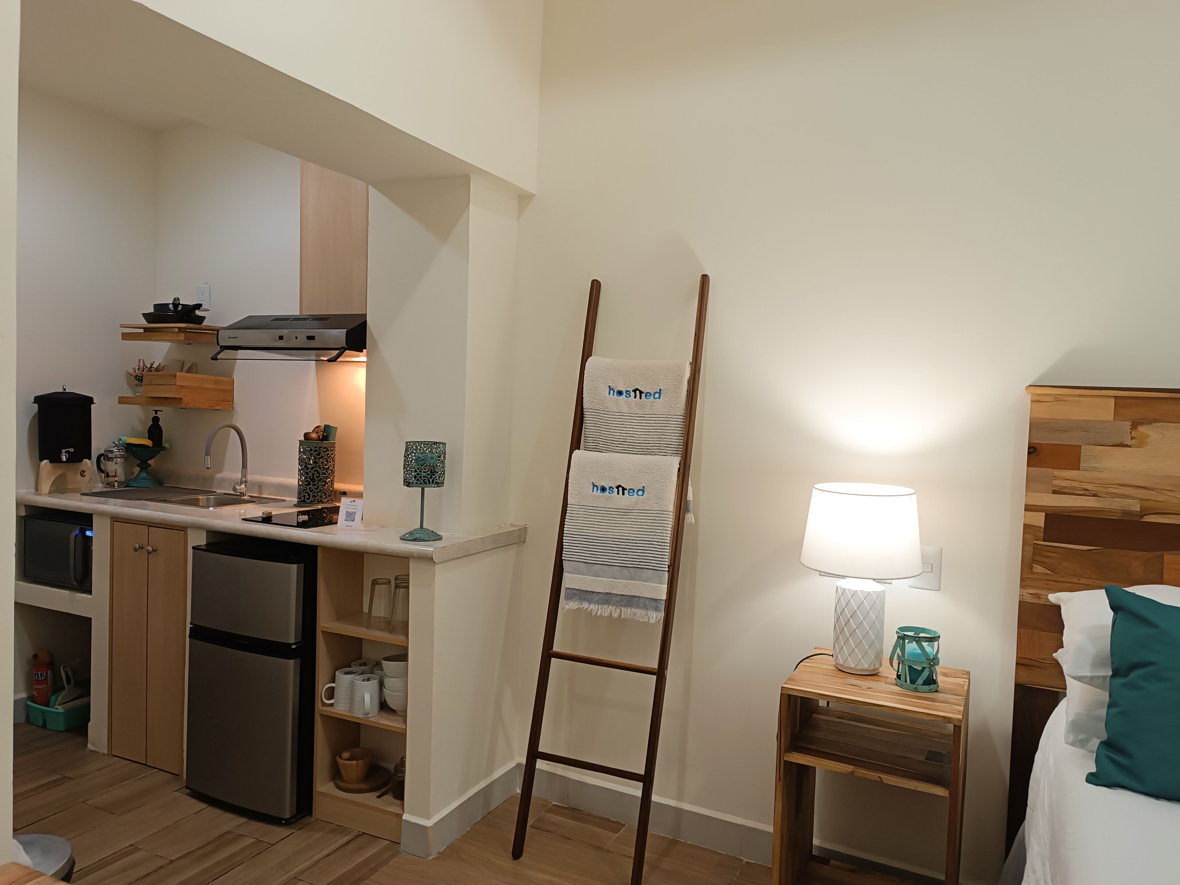 Una habitación de hotel compacta con kitchenette, una escalera con toallas, una mesita de noche con lámpara y una cama con almohadas color turquesa.