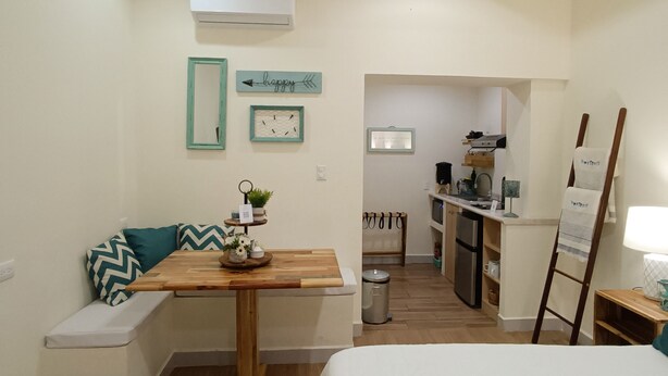 A compact bedroom with a bed, a small table with a plant, and a bench with pillows. A kitchenette is visible in the background. - Hostted (Flores)