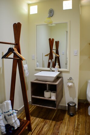 A bathroom with a wooden floor, a wooden hanger rack, a sink, a mirror, a toilet, and a trash can. - Hostted (Flores)