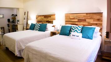 Two single beds with white linens and teal pillows, wooden headboards, bedside tables with lamps, and a kitchenette in the background.