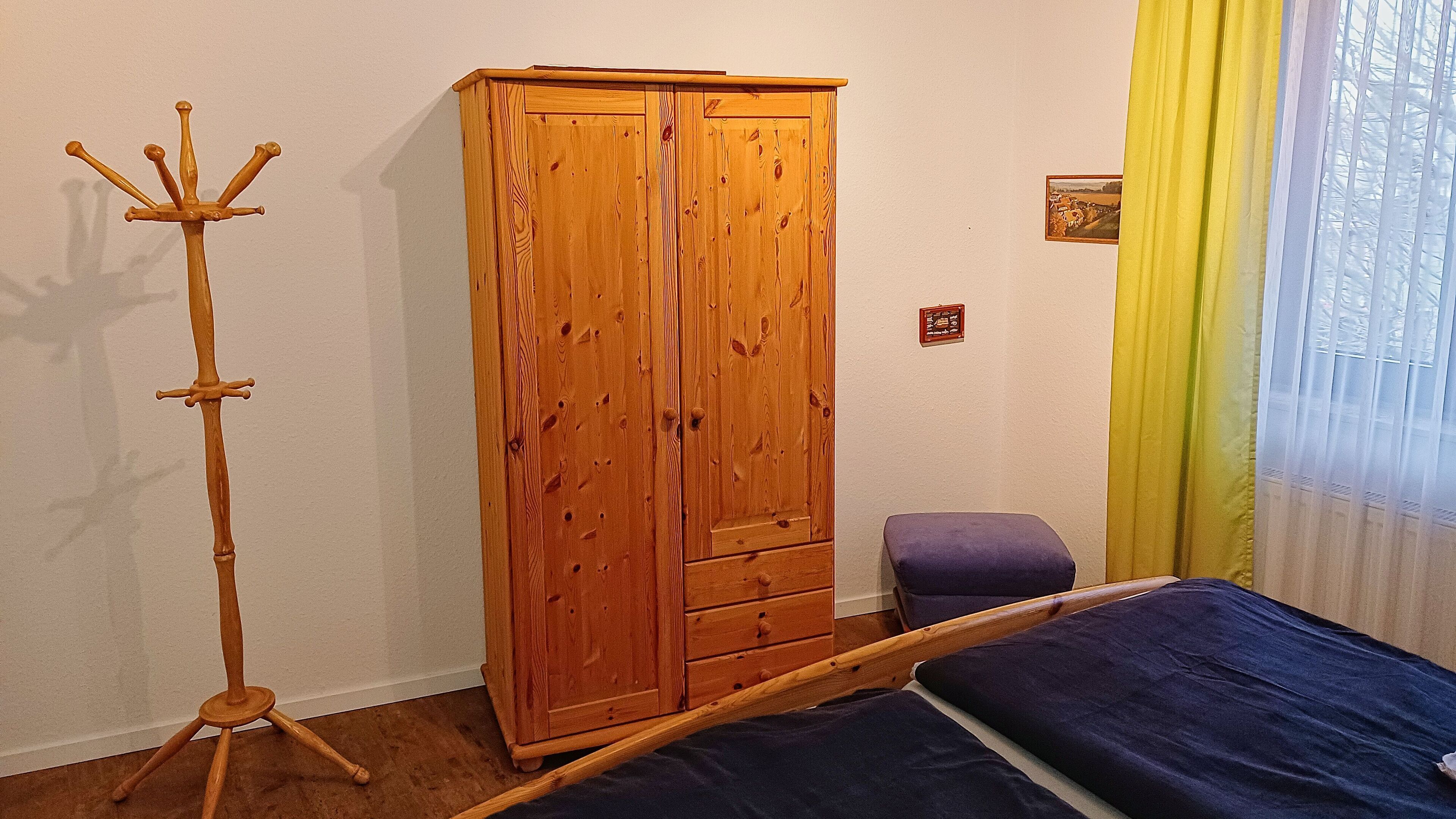 Un dormitorio con un armario de madera, un perchero de madera, una cama con colchón azul y un taburete pequeño morado.