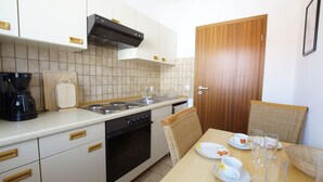 A compact kitchen with a stove, sink, and a coffee maker. There is a dining table with a chair and a door nearby.