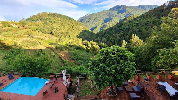 Zona de piscina con sillones reclinables y una terraza de madera, rodeada de vegetación exuberante y montañas al fondo.