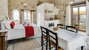 A rustic room with a stone wall, a bed with white bedding and red pillows, a dining table with chairs, and a kitchen area with a stove and cabinets. - Sanders Arsorama - Sweet 1-bdr Apt w/ Shared Patio (Arsos)