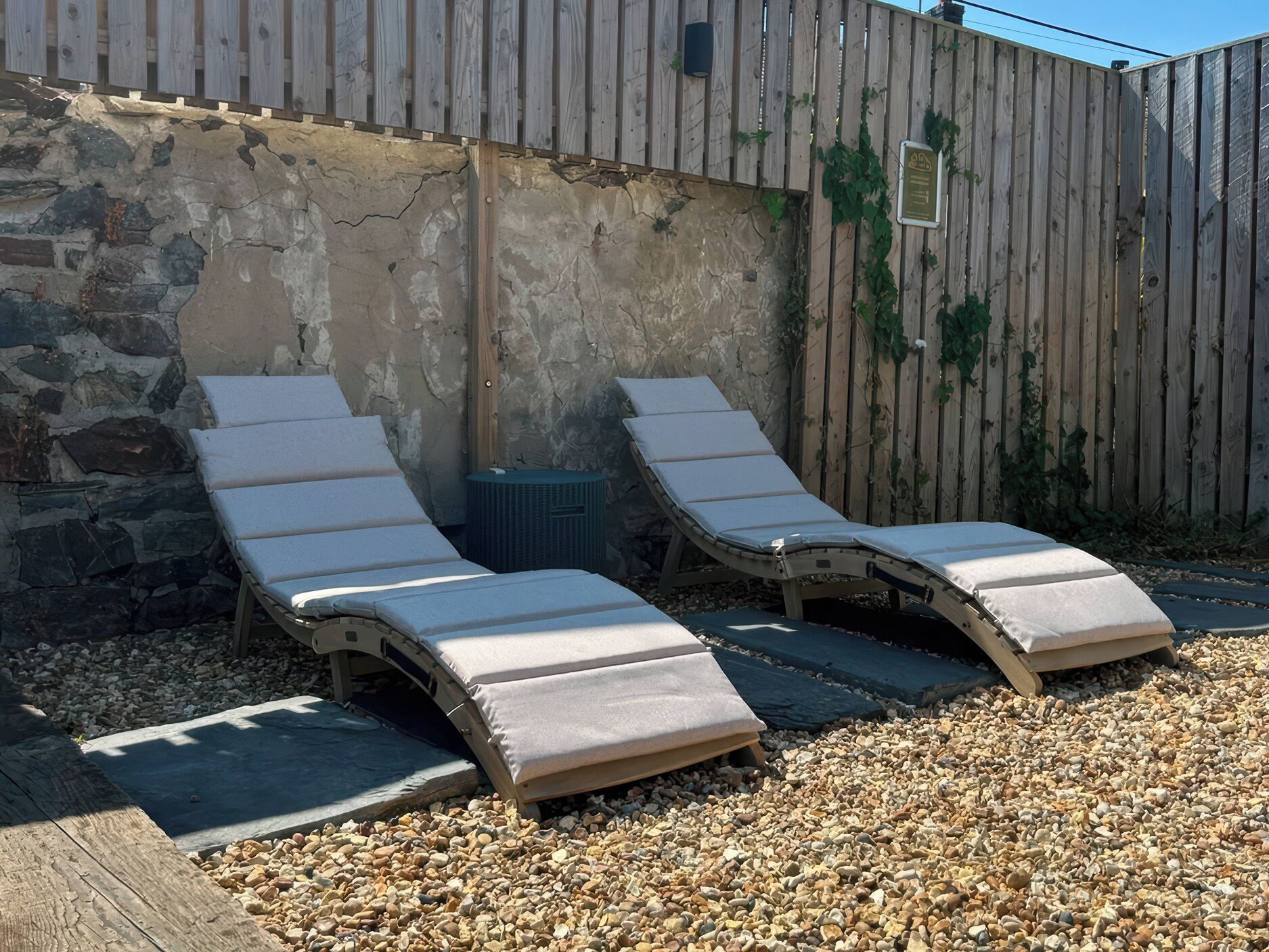Two sun loungers with cushions, a stone wall, and a wooden fence.