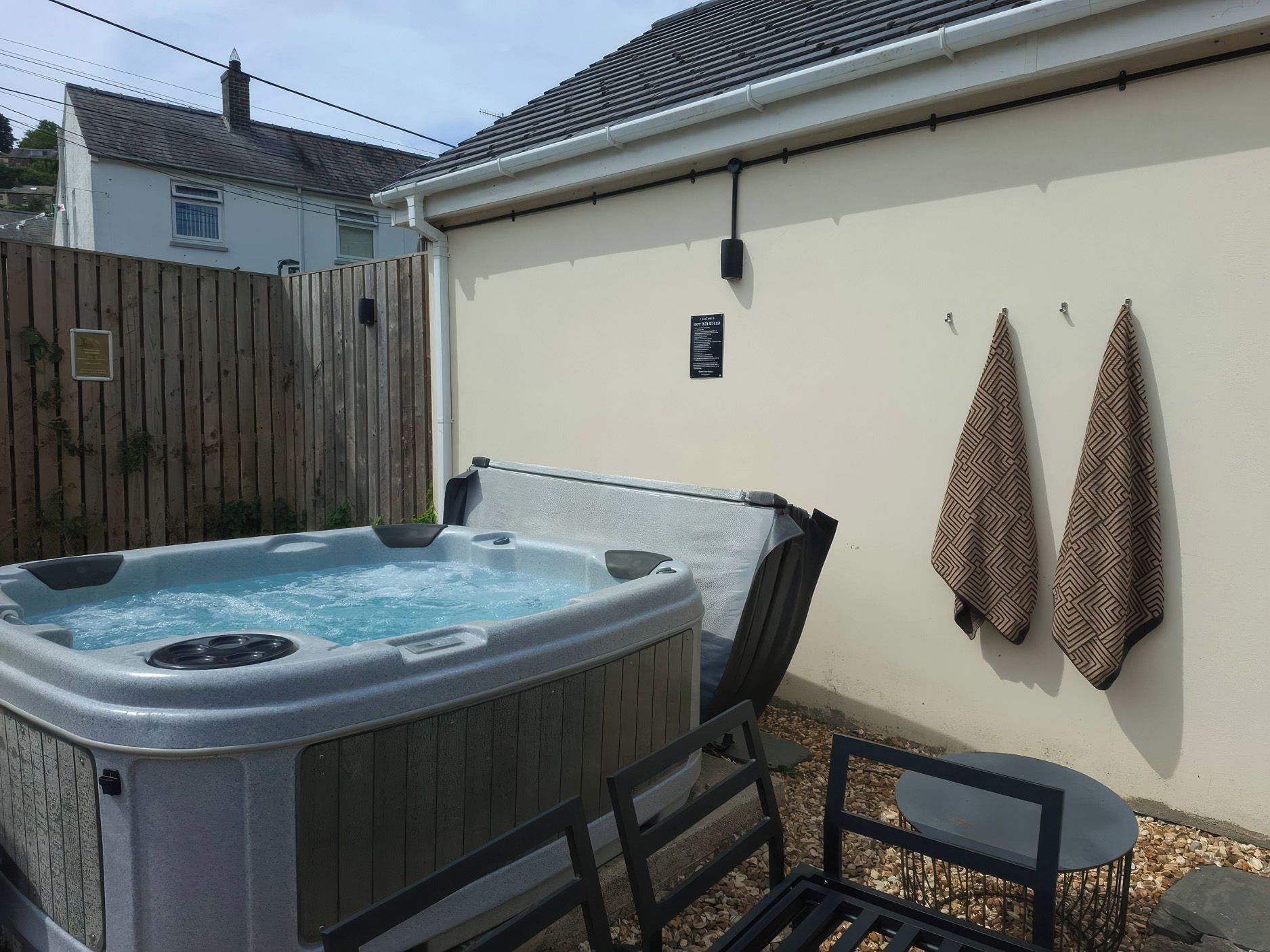 A hot tub in a backyard with a wooden fence, a wall-mounted towel rack, and a patio table.