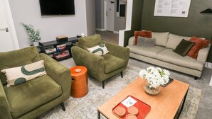 Ett modernt vardagsrum med två gröna fåtöljer, en grå soffa med mönstrade kuddar, en platt-TV som är monterad på väggen och ett träbord med diverse föremål på.