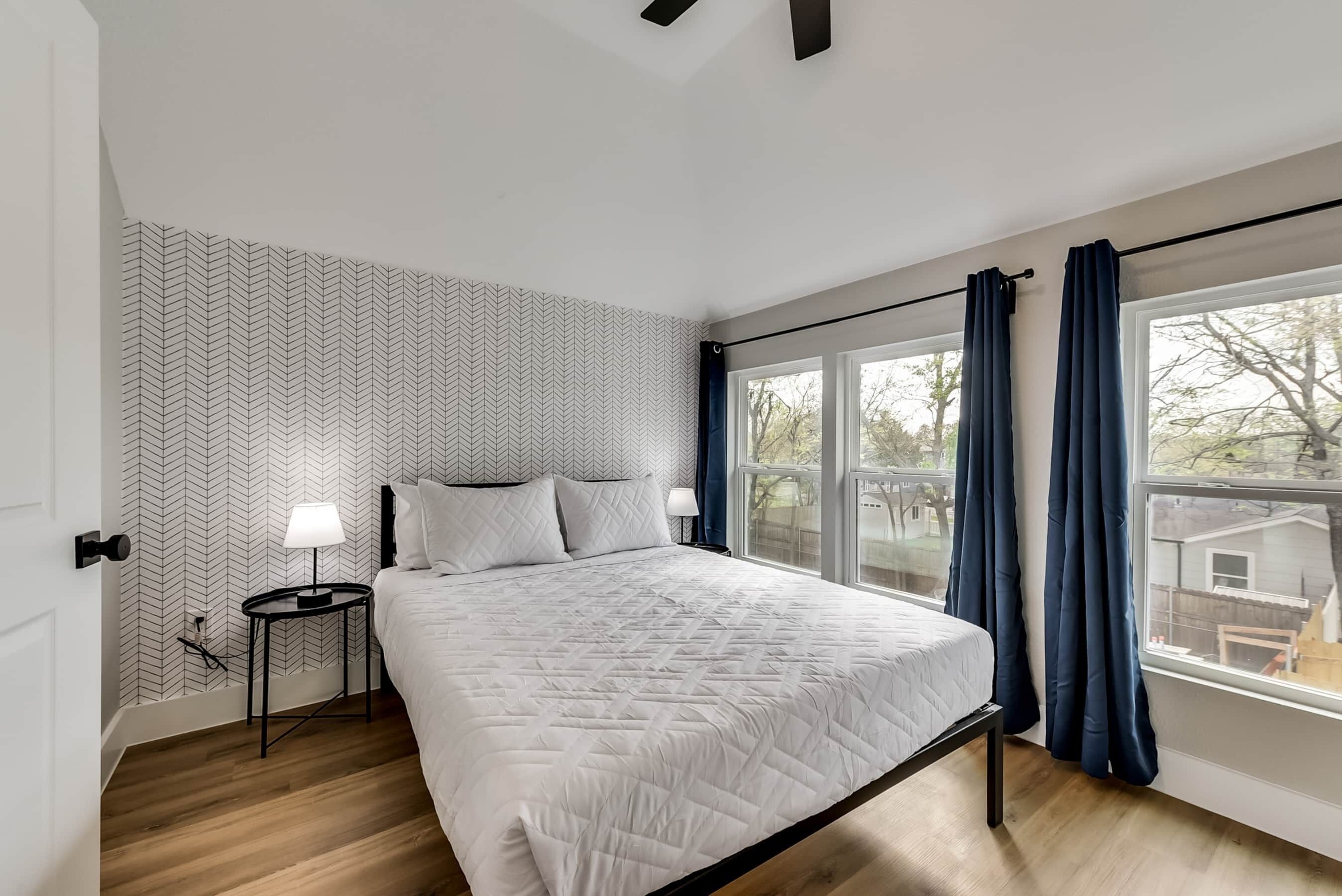 A bedroom with a bed, a nightstand, a window with curtains, and a patterned wall.