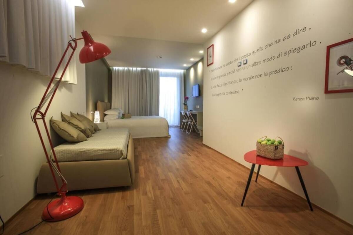 Une chambre d’hôtel moderne avec un plancher en bois, un canapé beige, un lampadaire rouge, une petite table rouge avec un panier, et un lit avec une tête de lit blanche.