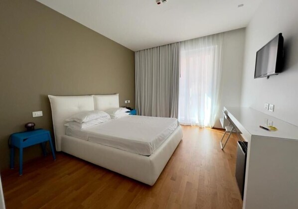 Una habitación de hotel moderna con una cama grande, un escritorio pequeño, un televisor montado en la pared y una ventana con cortinas.