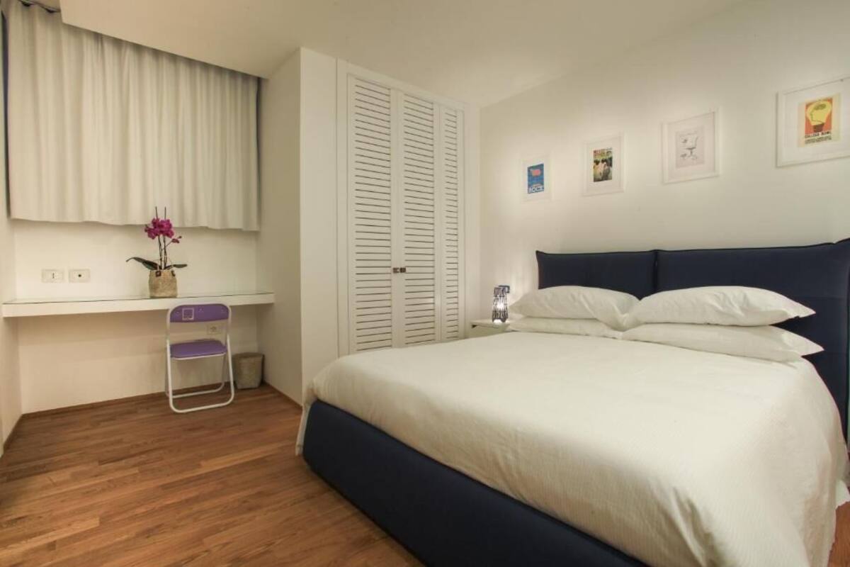 Une chambre à coucher comprenant un lit, une table de chevet, une commode, une fenêtre avec des rideaux et une petite table sur laquelle est posé un vase de fleurs.