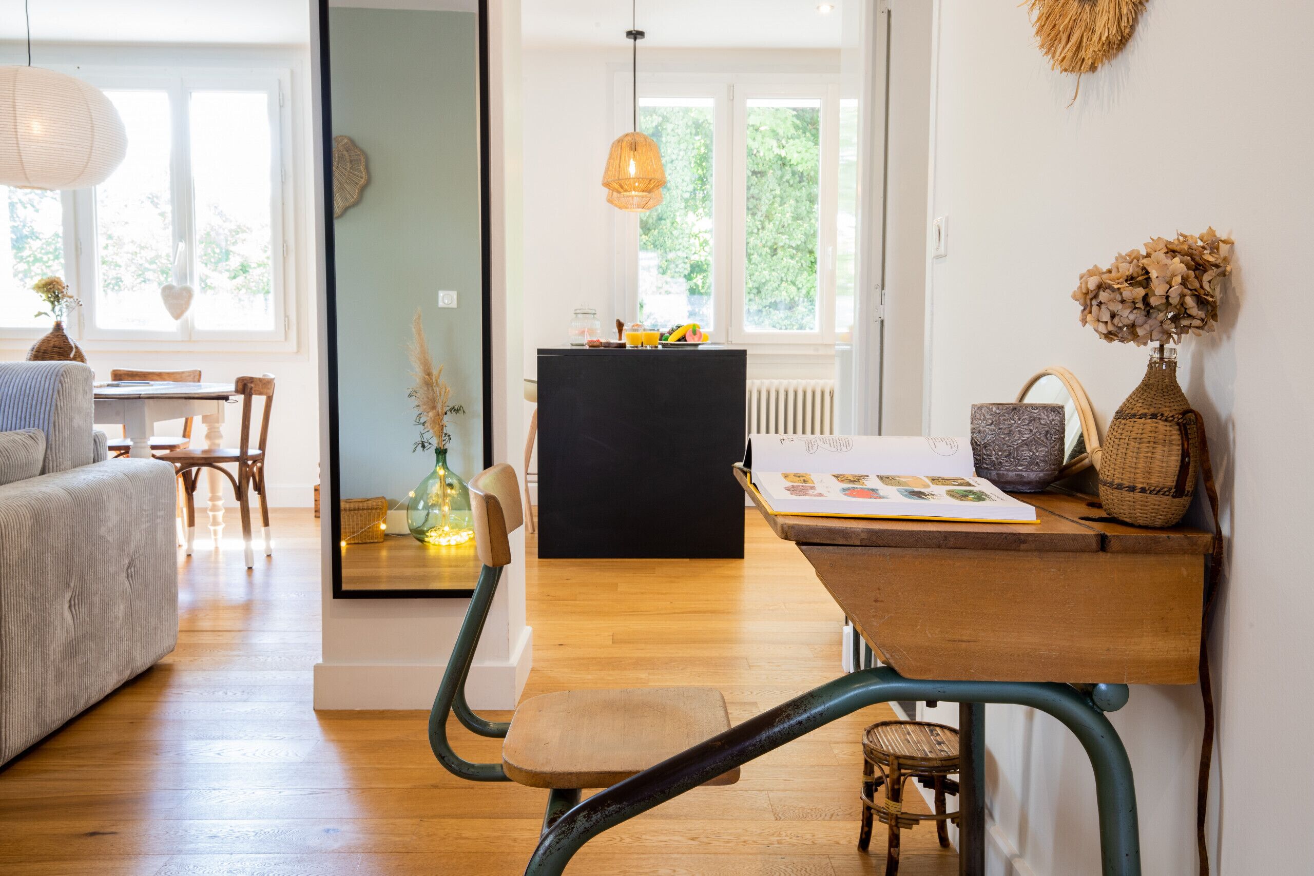 Ein gemütliches Wohnzimmer mit einer Couch, einem Holzesstisch und Stühlen. Es gibt eine schwarze Kommode mit Dekorationsgegenständen und ein Fenster mit Blick auf Grünflächen.